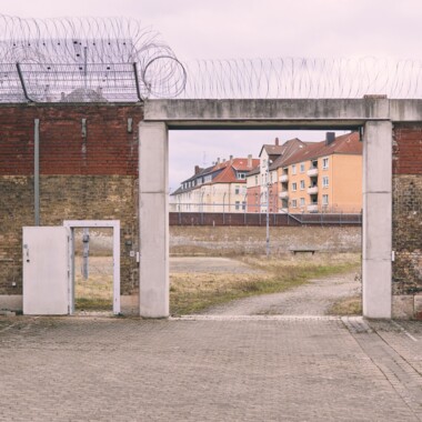 A view of the inner courtyard of Rennelberg Prison. A large gate in a high brick wall, behind which lies the former prison grounds. Barbed wire runs along the top of the wall; the surrounding residential buildings can be seen in the background.